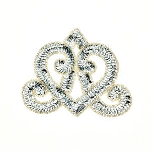 May include: A decorative silver sequin applique in a heart-shaped design. The applique features a central pointed element and two swirling heart shapes, all outlined with small, silver beads. The sequins catch the light, creating a shimmering effect.
