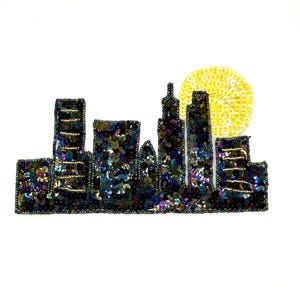 May include: A cityscape design, featuring buildings and a yellow sun, is crafted with sequins and beads. The buildings are dark with iridescent sequins, and the sun is a bright yellow circle. The design is set against a white background.