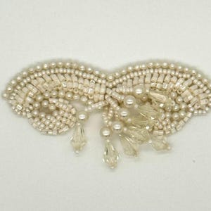 May include: Ivory-colored beaded applique with a symmetrical design. The applique features small, round beads and rectangular beads, with dangling teardrop-shaped crystal beads. The piece is likely for embellishing clothing or accessories.