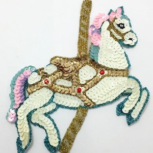 May include: A white, gold, and blue sequined carousel horse applique with pink and purple accents. The horse is standing on its hind legs with its head turned to the side. The horse has a gold saddle and a gold pole.