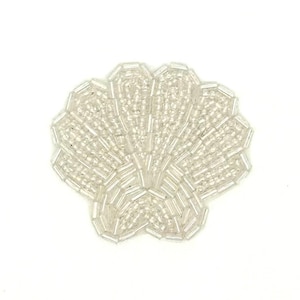 May include: A shell-shaped applique crafted from clear, translucent glass beads and elongated bugle beads. The design features a fan-like structure with radiating lines, creating a textured, shimmering surface that reflects light across the entire piece.