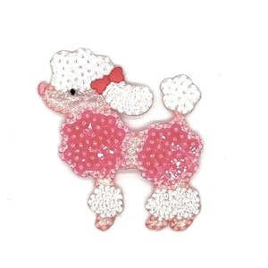 May include: An embroidered poodle patch featuring white thread for the head, tail, and leg puffs, and pink sequins covering the body and chest. A small red bow is attached to the poodle's head. The design is shaped like a standing dog.