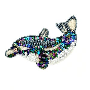 May include: A dolphin-shaped patch crafted with iridescent sequins in shades of blue, purple, and green. The design features a white sequin underbelly, a detailed fin, and a tail accented with silver beads, creating a shimmering, textured appearance.