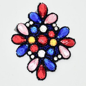 May include: A multi-colored rhinestone applique with a floral design. The applique features red, pink, blue, yellow, and white rhinestones, outlined with black beads. The design is set on a black mesh backing.