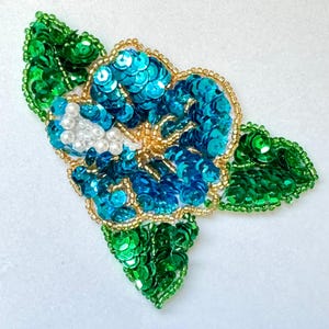 May include: A blue and green sequined flower with white pearl accents. The flower is made of sequins and beads and is perfect for adding a touch of sparkle to any project.