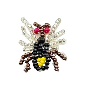 May include: A beaded insect brooch crafted with black, brown, yellow, and clear glass beads. The insect features two bright pink eyes, a black body with a yellow centre, brown legs, and translucent clear wings made of small cylindrical beads.