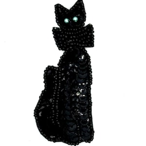 May include: A black cat brooch, made from sequins and beads. The cat is shown sitting, with bright blue eyes. The brooch is designed to be attached to clothing or accessories.