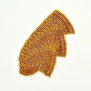 May include: A gold-colored, beaded applique in an abstract shape. The applique is made of small, rectangular beads arranged in a geometric pattern. The beads are a warm, metallic gold color, and the applique is set against a white background.