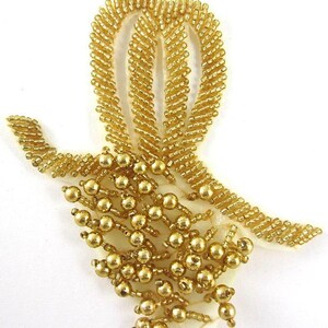 May include: A gold beaded applique with a bow-like design. The applique features a series of gold beads arranged in a decorative pattern, with a central loop and cascading beaded strands. The design is set on a cream-colored fabric.