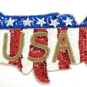 Peut inclure: Un écusson en forme des États-Unis, orné de paillettes rouges et blanches, de sequins bleus avec des étoiles blanches et les lettres "USA" en perles dorées.