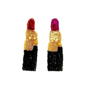 May include: A pair of beaded lipstick earrings. One earring has a red lipstick top, the other has a magenta top. The middle section is gold, and the bottom is black. The earrings are on a white background.