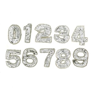 May include: A collection of silver sequined numbers from zero to nine, arranged on a white background. Each number is outlined with a silver border. The sequins give a sparkling, textured appearance to the numerals.