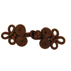 May include: A dark brown frog closure, a type of button, against a white background. The closure is made of tightly woven cord, featuring a series of loops and coiled elements. This decorative button is used for clothing and accessories.