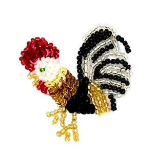 May include: A beaded rooster brooch featuring a vibrant red comb, a white head with a green eye, and a body crafted from gold, black, and white beads. The tail feathers are composed of alternating black and silver beads, with gold beaded legs.