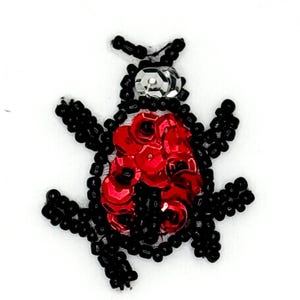 Peut inclure: Un écusson en forme de coccinelle composé de perles de rocaille noires pour les pattes et les antennes, d'une paillette argentée pour la tête et d'un amas de paillettes circulaires rouge vif formant le corps, sur un fond blanc uni.
