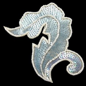 May include: A decorative applique shaped like a stylized feather or leaf, covered in iridescent light blue sequins. The edges are outlined with a border of small, white seed beads, creating a textured and reflective surface against the black background.