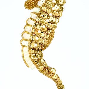 May include: A gold sequined seahorse, a marine-themed accessory. The seahorse is embellished with gold sequins, beads, and iridescent white accents. The seahorse is a decorative item, possibly a brooch or embellishment.