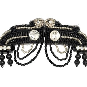Peut inclure: Une applique décorative symétrique composée de perles de rocaille noires et argentées, de perles tubes noires et de cristaux facettés transparents. Le motif inclut des rangées de perles noires pendantes et deux grands cristaux ronds sur filet.