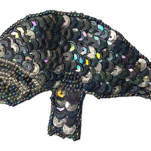 May include: A manatee-shaped patch covered in dark, iridescent sequins that shift between black, blue, and purple tones. The design features a rounded head, a single visible flipper, and a tapered tail, all meticulously hand-stitched with beads.