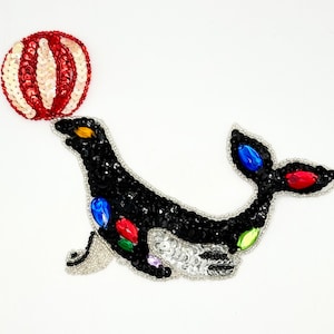 May include: A decorative patch shaped like a black seal balancing a red and white striped ball on its nose. The seal is embellished with black sequins, silver accents, and colorful faceted gems in blue, red, and green on its body and tail.