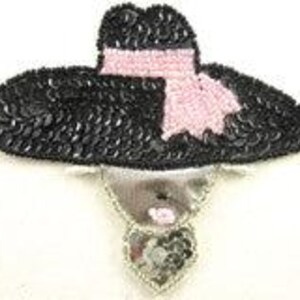 May include: A black sequined hat with a pink scarf and a silver face and heart-shaped pendant. The hat has a wide brim and a rounded crown. The pendant is embellished with silver beads. The overall design is a whimsical, decorative brooch.
