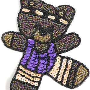 May include: A beaded and sequined raccoon applique with a purple vest and gold stripes. The applique is made of iridescent beads and sequins.
