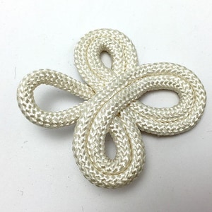 May include: A cream-colored decorative rope knot, resembling a four-leaf clover. The knot is made of tightly woven, thick rope, and is set against a white background. The rope has a textured appearance.