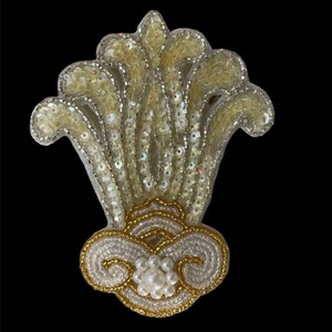 May include: A white and gold beaded floral design with a pearl center. The design is reminiscent of a stylized flower.