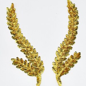 May include: A pair of gold sequin leaf-shaped patches. Each patch features a central stem with two branching segments covered in small, reflective gold sequins that create a textured, shimmering appearance against a plain white background.