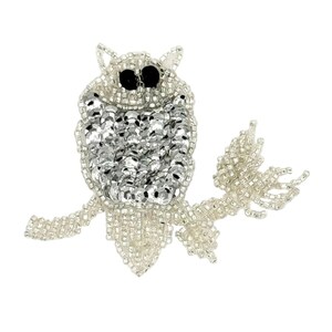 May include: A beaded owl brooch featuring silver seed beads and reflective silver sequins on the body. The owl has two prominent black bead eyes and is perched on a branch constructed from silver seed beads against a plain white background.