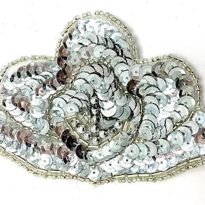 May include: A silver sequin applique in a floral design. The applique is made of numerous small, round, shiny silver sequins, with a beaded edge. The sequins are arranged in a layered pattern, creating a textured surface. This decorative applique is suitable for embellishing clothing or accessories.