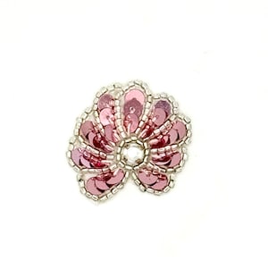 May include: A floral-shaped patch featuring pink circular sequins arranged in a petal pattern. The edges of each petal are outlined with small silver beads, and a single clear faceted crystal is positioned at the center of the flower design.