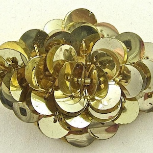 May include: A cluster of gold-colored sequins arranged in a flower shape. The sequins are overlapping and create a three-dimensional effect.