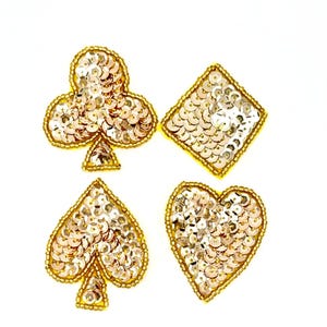 Peut inclure: Quatre écussons de jeu de cartes : un trèfle, un carreau, un pique et un cœur. Chaque écusson est orné de paillettes argentées et bordé de perles dorées. Les écussons sont posés sur un fond blanc.