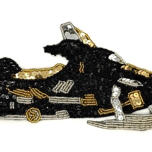 May include: A decorative patch shaped like a snowmobile, crafted with black, gold, and silver beads and sequins. The design features a black body with gold and silver accents, including a silver beaded ski at the front and detailed beadwork.