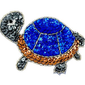 May include: A turtle-shaped patch embellished with sequins. The head and limbs are covered in dark gray sequins, while the shell features a central section of bright blue sequins surrounded by a border of metallic copper-colored sequins.