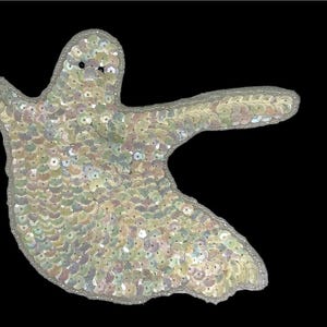 May include: A white ghost-shaped applique made of iridescent sequins. The ghost has a rounded head and a long, flowing body.