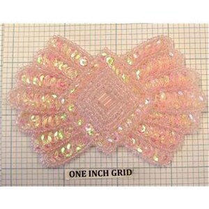 May include: A pink sequined bow-shaped embellishment with a central square design. The bow is approximately 3 inches wide, with iridescent sequins and beads. The background features a one-inch grid for scale.