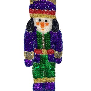 May include: A beaded nutcracker ornament with a purple, green, gold, and red sequined design. The nutcracker has a crown, a white face with black hair, and a red sash.