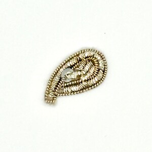 May include: A teardrop-shaped, silver-colored embellishment with a central, clear, round gem. The embellishment is made of tightly woven, metallic threads, creating a textured surface. The design is set against a plain white background.