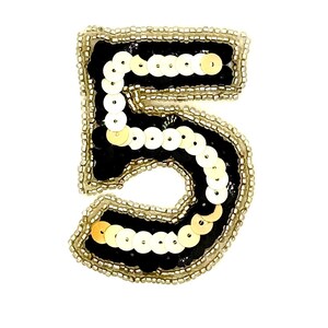 May include: A decorative patch in the shape of the number five, featuring a black felt base adorned with rows of shimmering gold sequins and outlined with a border of small, metallic gold seed beads, creating a textured and reflective surface.