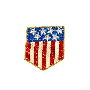 May include: A patriotic shield-shaped patch featuring the American flag design. The top section has a blue background with white star sequins, while the bottom has red and white striped sequins. The border is outlined with gold beads.