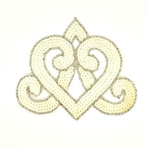 May include: Ivory-colored sequined applique in a heart-shaped design with a silver beaded outline. The applique features a central diamond shape and two symmetrical swirls. The design is on a white background.