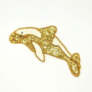 May include: A dolphin-shaped patch embellished with gold and white sequins. The design features a gold sequined body and tail with white sequined accents on the head, pectoral fin, and dorsal fin, all outlined with gold beads on a white background.