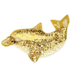 May include: A dolphin-shaped brooch crafted with gold sequins and small beads. The design features a layered texture of metallic gold sequins covering the body, with fine beadwork outlining the fins, tail, and snout against a white base.