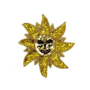 May include: A gold sequined sun-shaped appliqué with a smiling face. The sun has radiating rays and a central face with black and brown details. The sequins are gold and the face has a happy expression. The appliqué is on a white background.