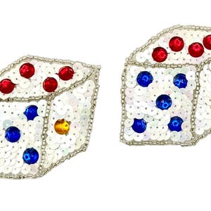 Peut inclure: Deux dés recouverts de paillettes blanches et bordés de perles argentées. Les dés ont des points rouges, bleus et jaunes en forme de bijoux représentant les chiffres. Les dés sont posés sur un fond blanc.
