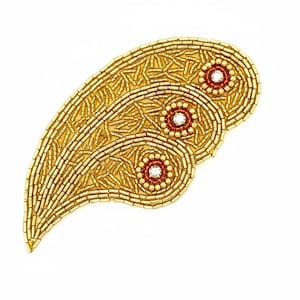 May include: Gold-colored beaded paisley-shaped embellishment. The design features three circular accents with red centers and clear crystal-like embellishments. The paisley is made of small, gold-colored beads.