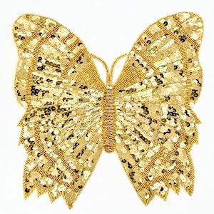 May include: A gold sequined butterfly with intricate beadwork. The butterfly's wings are adorned with gold sequins and beads, creating a shimmering effect. The body is a vertical line of gold beads. The butterfly is a decorative item.