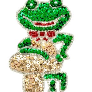 May include: A beaded patch featuring a green frog with a red bow tie, sitting on a gold sequined mushroom. The frog is crafted with bright green sequins, while the mushroom cap and stem are covered in shimmering gold sequins and silver beads.